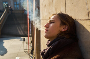 Young casual handsome guy with brown hair exhaling the white smoke of a cigarette on a sunny winters day, while leaning on a wall in Berlin city.
