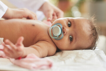 Close up portrait of cute African-American baby sucking on pacifier and looking at camera while lying on changing table with mother dressing him, copy space