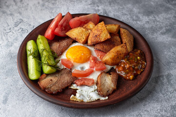 Homemade breakfas on a platet, salted cucumbers, fried potatoes, fried egg, bacon, tomatoes and homemade sauce, on white concrete