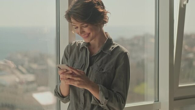 Happy young pretty woman using smartphone while standing near the window at home