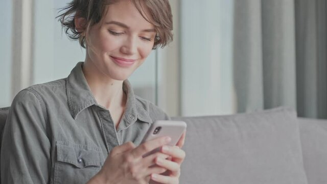 Happy young pretty woman writing message on smartphone while sitting on sofa at home
