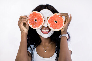 Young cheerful African woman with white clay mud facial mask holds big grapefruits slices, covering her eyes, and smiles to camera. Healthy diet, skincare, facial treatment, cosmetology concept.