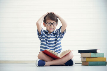 A little Caucasian boy wear eye glasses reading a book and sitting on the floor at home. He took...