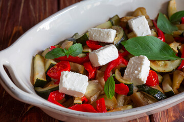 Ratatouille, backed vegetable with feta cheese and fresh basil in white oven pan on wooden brown background.