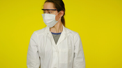 Young doctor in a protective medical mask, glasses and a white coat. Girl nurse on a yellow background. Coronavirus, flu