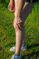 A young woman standing on green grass holds a damaged knee. Sprain or cramps Overstrain an injured person while training or running outdoors.