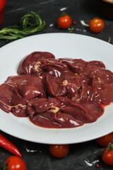 Chicken ventricles in a in a white plate, fresh rosemary, fresh cherry tomatoes, fresh chili peppers, garlic, lime and sea salt on a black background.