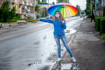 Caucasian girl in a protective mask hails taxi on an empty street, stands with an umbrella in spring rain and waits for car. Safety and social distance during coronavirus pandemic. Copy space.