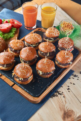 Juicy mini burgers with onion rings, jalapeno pepper, cucumbers, lettuce, cheese on a black stone background. Orange and carrot juce