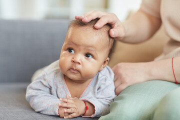 Front view portrait of cute mixed-race baby crawling on couch with unrecognizable Caucasian mother laying hand on head, copy space