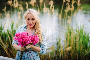 Portrait of a young blonde woman with a bouquet of pink peonies .. A girl in a blue dress and a hat with flowers is resting on the beach of a pond. The concept of outdoor recreation. Summer holidays.
