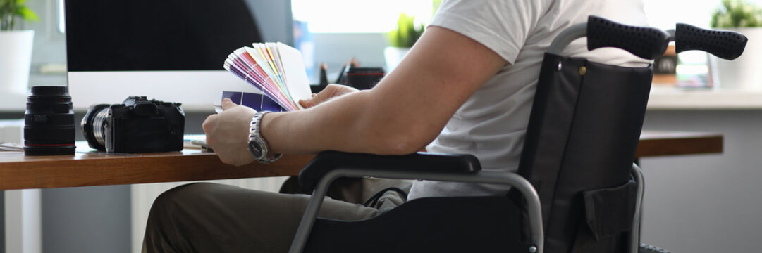 Close-up of professional designer working in office holding colourful palette. Male sitting in wheelchair. Photocamera and laptop on desktop. People with disability concept