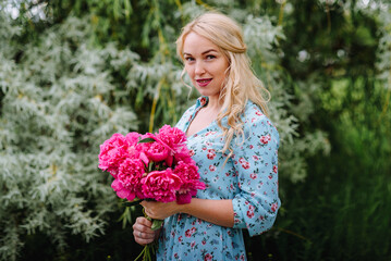 Portrait of a young blonde woman with a bouquet of pink peonies .. A girl in a blue dress and a hat with flowers is resting in a green park. The concept of outdoor recreation. Summer holidays.