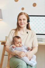 Vertical portrait of adult working mother holding baby and smiling at camera while posing next to...