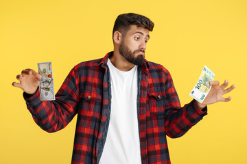 Confused bearded hipster young man in plaid shirt holding dollar and euro banknote over yellow background.