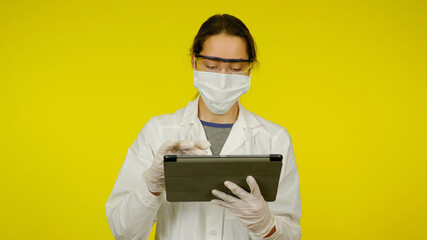 Young doctor in a medical mask, goggles and latex gloves looks at the tablet. Girl in a white coat on yellow background holds tablet in hands and looks at patient information. Coronavirus, flu