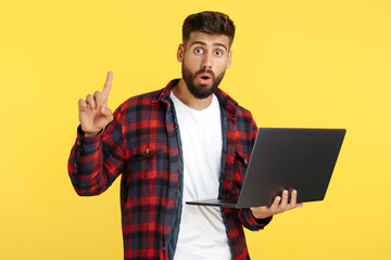 Excited bearded hipster young man in plaid shirt holding notebook show finger up over yellow...