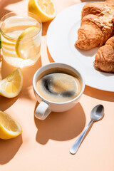coffee cup with teaspoon, croissants and glass of water with lemon for breakfast on beige table