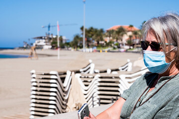 Fototapeta na wymiar Senior adult woman wearing a protective mask looks at deserted beach with closed umbrellas and beach beds - lockdown due to the Covid-19 coronavirus.