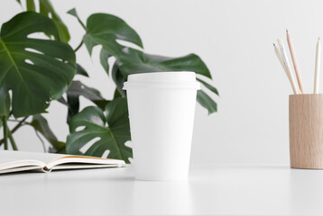 Coffee paper cup mockup  with a notebook, workspace accessories and a monstera plant on a white table.