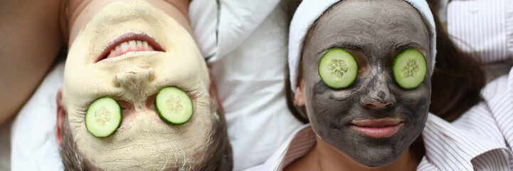 Top view of man and woman in face masks laying on bed. Moisturizing treatment and cucumbers on eyes. Beauty procedure for couple. Skincare and cosmetology concept