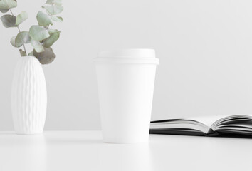 Coffee paper cup mockup with eucalyptus in a vase and a notebook on a white table.