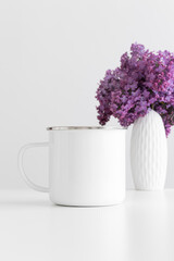 Enamel mug mockup with a bouquet of lilac in a vase on a white table.