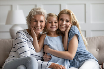 Three different age and generation relatives beautiful females sitting on sofa hugging posing looking at camera. Women or Mother Day holiday celebration, multigenerational family bond and ties concept - Powered by Adobe