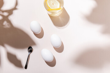 top view of white boiled chicken eggs, water with lemon and teaspoon on grey table with shadows
