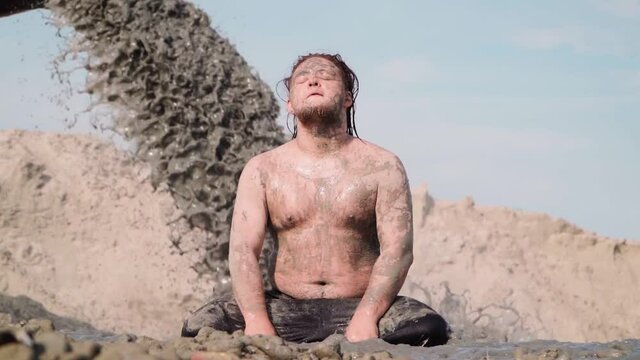 Funny Fat Man with Long Hair Sits in the Mud. Against the background pouring Black Dirty Water from the Pipe. Environmental pollution. Slow motion