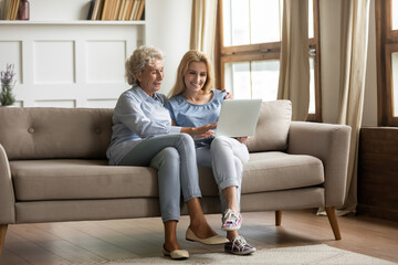 Obraz premium Mature mother grown up daughter sit on couch in living room hold on lap pc looking at screen watching movie film, using device make video call, shopping via e-commerce website, girl teach mom concept