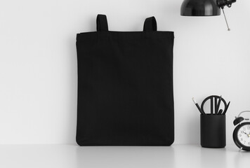 Black tote bag mockup with workspace accessories on a white table.