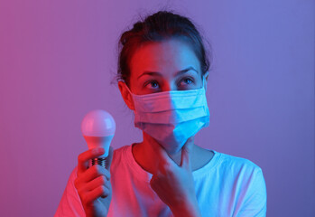 Pensive woman with medical face mask holds light bulb in red-blue neon light