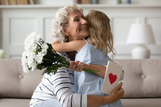 Sincere old grandma hugs small attentive granddaughter express her gratitude for received surprise at birthday or 8-march Woman Day. Grandchild makes surprise for granny gave her flowers and postcard
