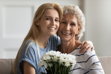 Head shot grown up daughter prepare for aged mom flowers, family cuddling seated on couch looking at camera. Mothers Day or International Women Day 8-march or anniversary holidays celebration concept