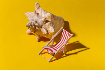 Mini beach deck chair and seashell on yellow background with shadow. Sunlight. Symbol of beach holidays, resort.  Relax, Summer minimal concept.