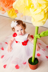 Cute toddler girl in white and red dress sits near huge yellow decorative flower.