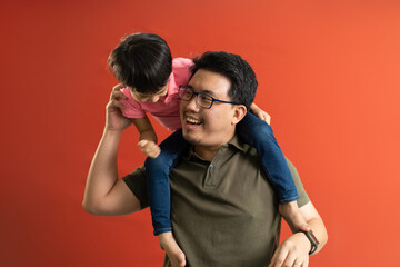 Asian father giving son ride on the back with a red background. Portrait of a happy father giving son piggyback a ride on his shoulders and looking up.