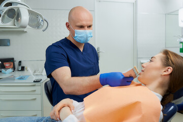 A bald middle-aged dentist chooses tooth shade for a pretty female patient before a whitening procedure in a dentist's office. A doctor is matching a tooth color to a patient before treatment.