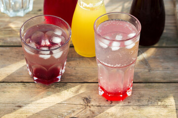 Fototapeta na wymiar Strawberry, cherry and rhubarb syrups and glasses with water on a wooden table in the garden.