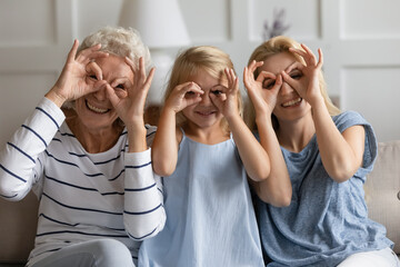 Multigenerational women sit on couch fooling around do funny faces making binoculars with fingers...
