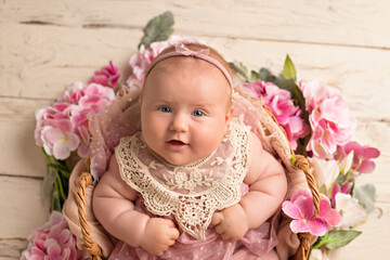 Happy smiling baby girl in a pink dress lies on her back in a flower basket on a white wooden background