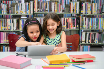 Two little happy cute girls playing on a tablet PC computing device in library at school. Education and self learning wireless technology concept. People lifestyles and friendship. Preschool children