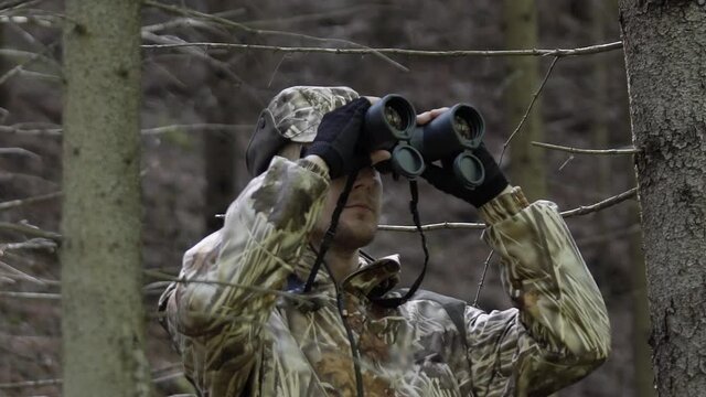 Hunter looks through binoculars. Man in comfortable camouflage clothes hunter outdoor in forest hunting alone.
