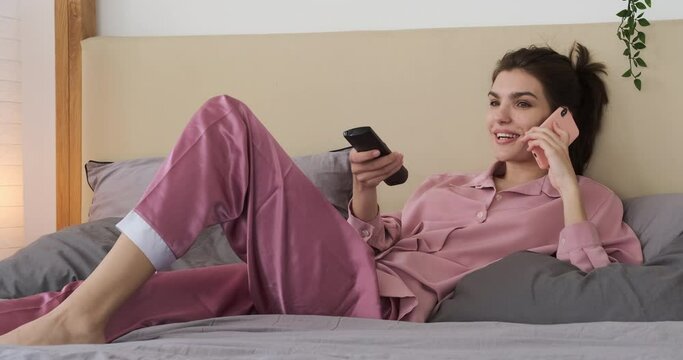 Woman in pajamas talking on mobile phone while watching tv and relaxing on bed at home