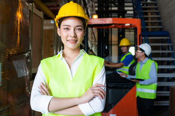 Portrait of Asian warehouse woman worker smile with arms crossed, supervisor talking with forklift driver at the background. export import or logistics service business. employee and labor factory