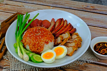 Barbecued red pork, crispy pork belly, Chinese sausage and boiled egg with rice topped with red sauce served with spring onion and cucumber slices having pickled chilli aside on wooden table.