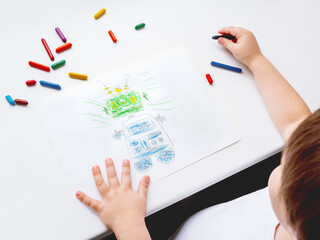 Toddler draws funny robot. Kid uses wax crayons. Top view on child's hands and pencils on white background.