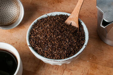 Bowl of roasted and ground barley on wood with a moka pot and a cup of barley coffee