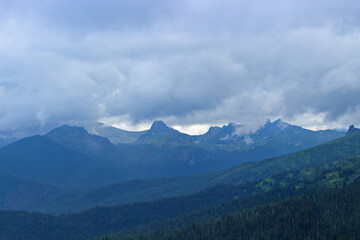 Fototapeta premium Blurry view to distant conifer forest and foggy mountains. Dark siberian taiga in haze. Low clouds over a mountain range. Landscape of hills covered in forests. Nature Park Ergaki, Russia, Siberia.